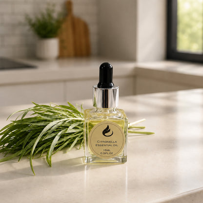 A bottle of Citronella Essential Oil with a black dropper cap and silver collar, placed on a light countertop with fresh green citronella leaves beside it. The background shows a softly blurred kitchen interior.